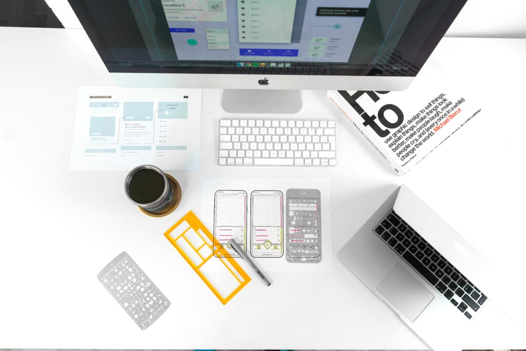 Website design layouts displayed on desk while computer monitor displays design notes